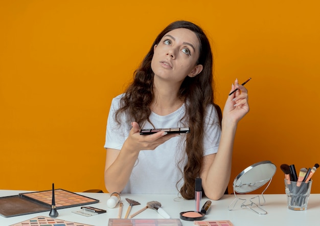 Chica guapa joven pensativa sentada en la mesa de maquillaje con herramientas de maquillaje con paleta de sombra de ojos y pincel mirando hacia arriba