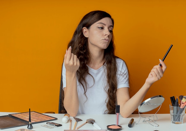 Chica guapa joven pensativa sentada en la mesa de maquillaje con herramientas de maquillaje sosteniendo y mirando el delineador de ojos manteniendo la mano en el aire aislado sobre fondo naranja