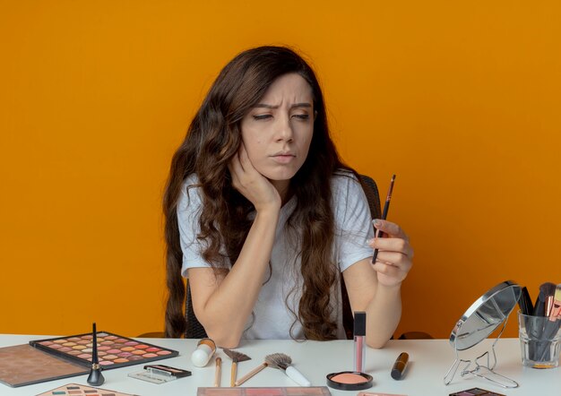 Chica guapa joven pensativa sentada en la mesa de maquillaje con herramientas de maquillaje sosteniendo y mirando el pincel de sombra de ojos