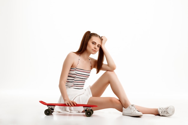 Chica guapa joven posando con patineta sobre pared blanca