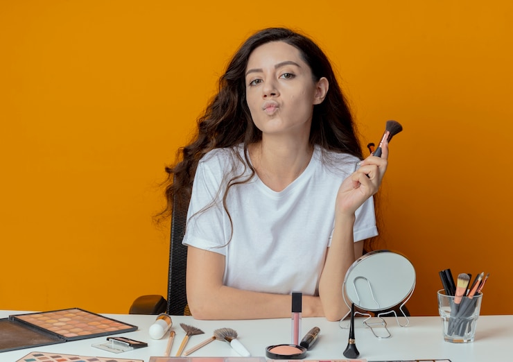 Foto gratuito chica guapa joven segura sentada en la mesa de maquillaje con herramientas de maquillaje sosteniendo pinceles de maquillaje aislado sobre fondo naranja