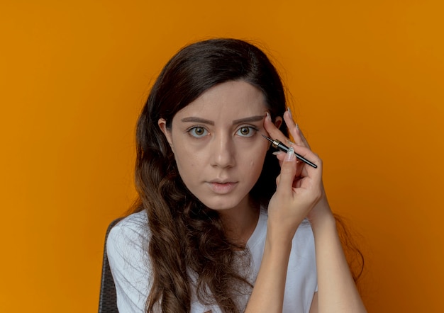 Chica guapa joven sentada en la mesa de maquillaje con herramientas de maquillaje aplicando delineador de ojos y tocando la cara mirando a cámara aislada sobre fondo naranja