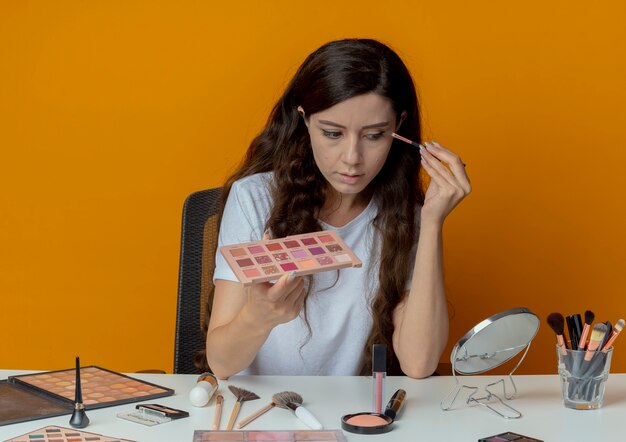 Chica guapa joven sentada en la mesa de maquillaje con herramientas de maquillaje mirando en el espejo y aplicando sombra de ojos aislado sobre fondo naranja