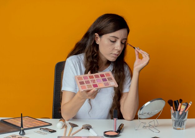 Chica guapa joven sentada en la mesa de maquillaje con herramientas de maquillaje mirando en el espejo y aplicando sombra de ojos con los ojos cerrados