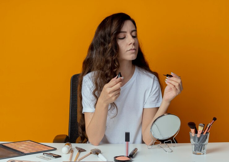 Foto gratuito chica guapa joven sentada en la mesa de maquillaje con herramientas de maquillaje sosteniendo y mirando delineador de ojos aislado sobre fondo naranja