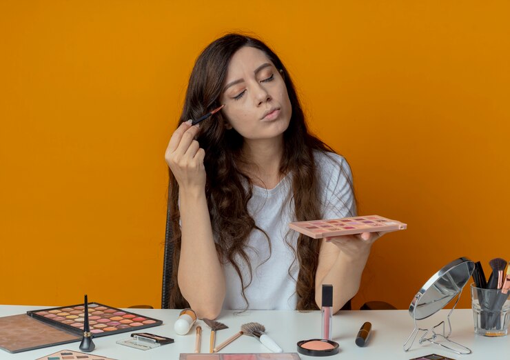 Foto gratuito chica guapa joven sentada en la mesa de maquillaje con herramientas de maquillaje sosteniendo la paleta de sombras y aplicando sombra de ojos con pincel con los ojos cerrados aislados sobre fondo naranja