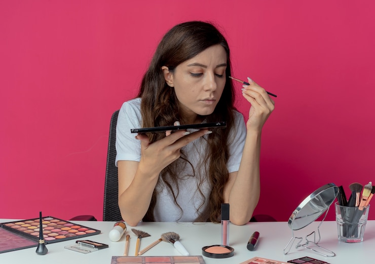 Foto gratuito chica guapa joven sentada en la mesa de maquillaje con herramientas de maquillaje sosteniendo la paleta de sombras de ojos mirando en el espejo y aplicando sombra de ojos con un ojo cerrado aislado sobre fondo carmesí