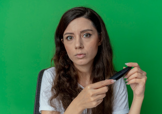 Chica guapa joven sentada en la mesa de maquillaje con herramientas de maquillaje sosteniendo rímel mirando a cámara aislada sobre fondo verde