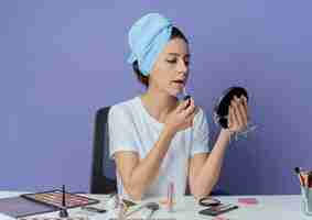 Foto gratuita chica guapa joven sentada en la mesa de maquillaje con herramientas de maquillaje y con una toalla de baño en la cabeza sosteniendo y mirando el espejo y aplicando brillo de labios aislado sobre fondo púrpura