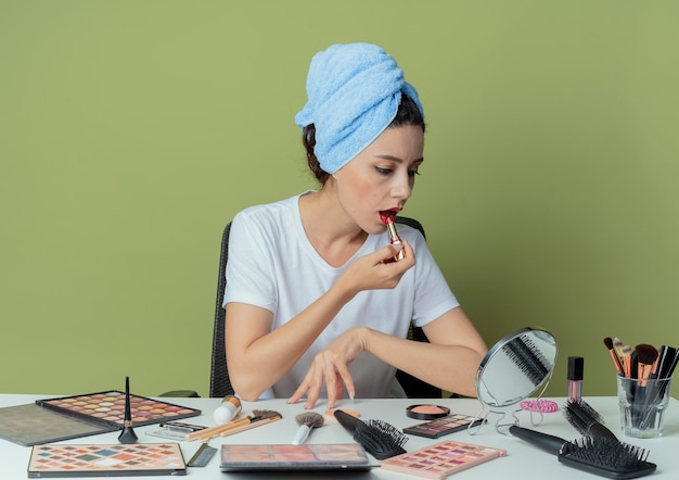 Chica guapa joven sentada en la mesa de maquillaje con herramientas de maquillaje y con una toalla en la cabeza mirando al espejo y poniéndose lápiz labial rojo y tocando la mesa en el espacio verde oliva