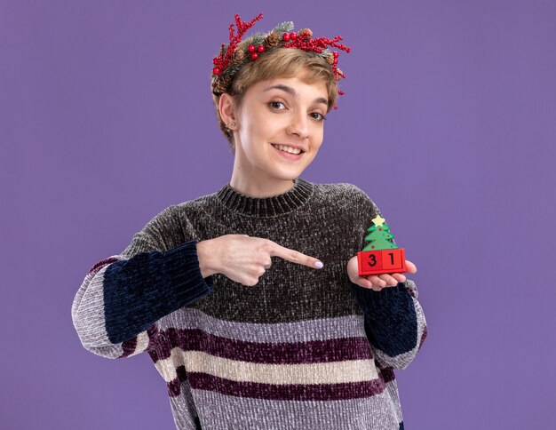 Chica guapa joven sonriente con corona de cabeza de Navidad sosteniendo el juguete del árbol de Navidad con fecha apuntando a él mirando a cámara aislada sobre fondo púrpura
