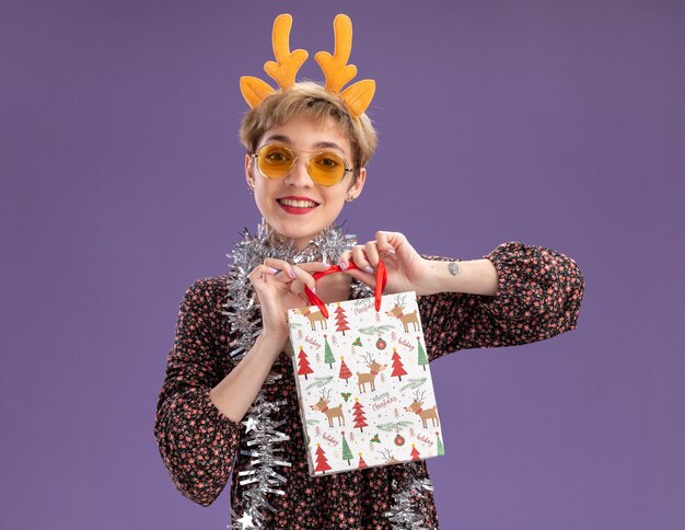 Chica guapa joven sonriente con diadema de cuernos de reno y guirnalda de oropel alrededor del cuello con gafas sosteniendo una bolsa de regalo de Navidad mirando a cámara aislada sobre fondo púrpura