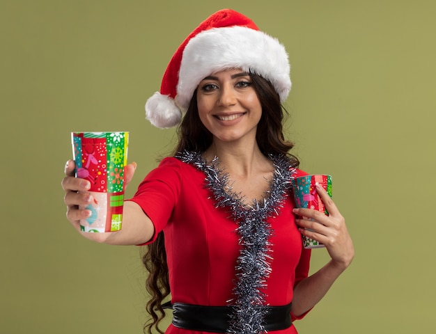 Chica guapa joven sonriente con gorro de Papá Noel y guirnalda de oropel alrededor del cuello sosteniendo tazas de café de Navidad estirando uno aislado en la pared verde oliva