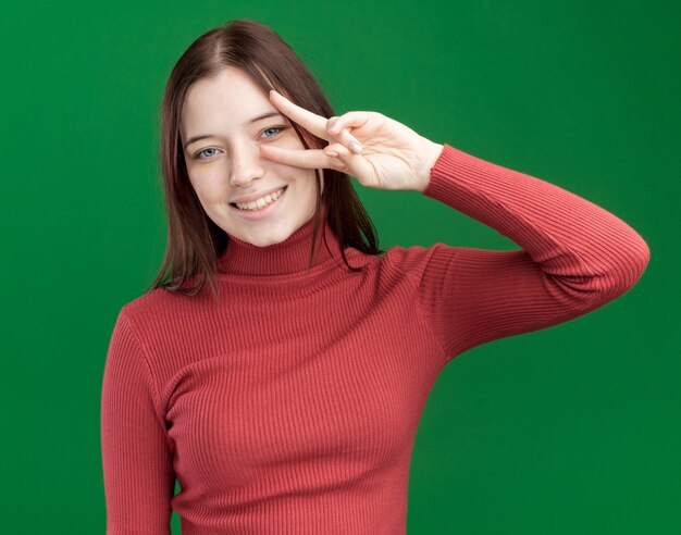 Chica guapa joven sonriente que muestra el símbolo v-sign cerca del ojo aislado en la pared verde