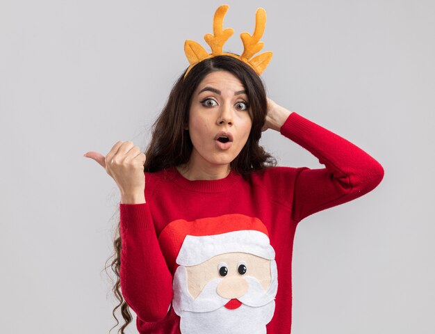 Chica guapa joven sorprendida con diadema de cuernos de reno y suéter de santa claus mirando manteniendo la mano en la cabeza apuntando al lado