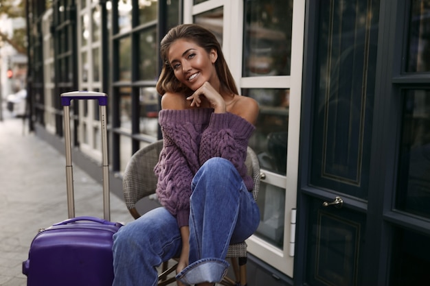 Chica guapa con la maleta sentada en la silla cerca del café, en el jersey morado, jeans, maquillaje, peinado, emociones, otoño, rubia, feliz, tejida, sonriente, coqueteando