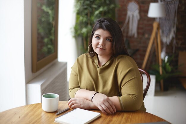 Chica guapa con mejillas regordetas y cuerpo curvilíneo haciendo tarea en casa sentado espacio de coworking con diario vacío en blanco y taza. Elegante mujer de talla grande bebiendo capuchino, tomando notas en el cuaderno