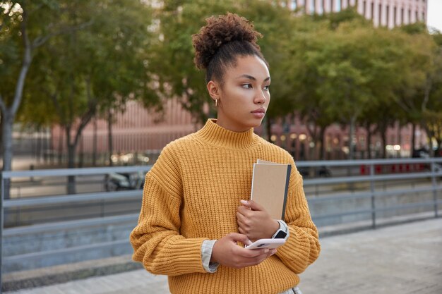 Chica guapa milenaria con expresión pensativa utiliza un teléfono inteligente digital y un cuaderno de bocetos educativo pasea durante el día en la ciudad viste un jersey de punto amarillo enfocado