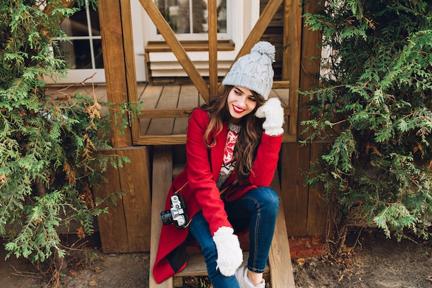 Chica guapa morena con abrigo rojo, gorro de punto y guantes blancos sentado en escaleras de madera al aire libre. Ella tiene el pelo largo, sonriendo a los lados.