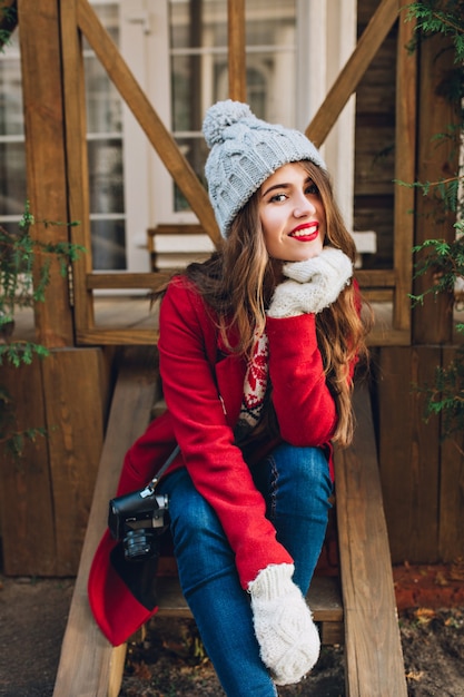 Chica guapa morena con abrigo rojo, gorro de punto y guantes blancos sentado en escaleras de madera al aire libre. Ella tiene el pelo largo, sonriendo.