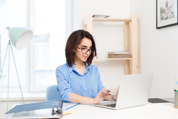 Una chica guapa morena con una camisa azul sentada a la mesa en la oficina. Ella está escribiendo en la computadora portátil y se ve feliz.