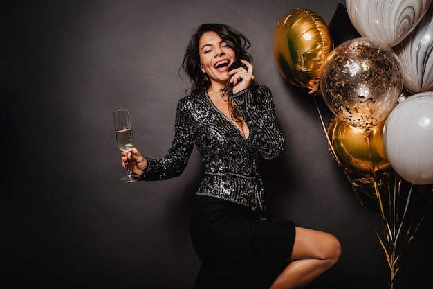 Chica guapa morena con copa de vino bailando sobre fondo negro Elegante modelo femenino disfrutando de la fiesta de cumpleaños