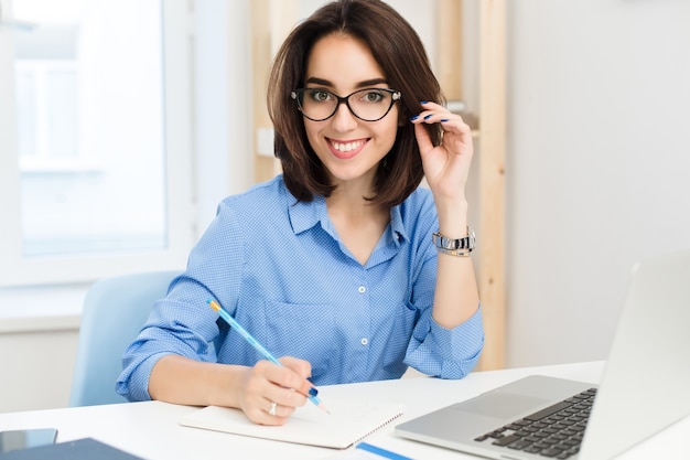 Una chica guapa morena está sentada a la mesa en la oficina. Viste camisa azul y gafas negras. Ella está escribiendo y sonriendo a la cámara.