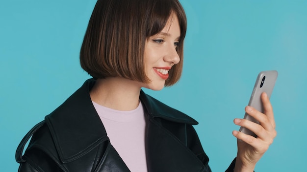 Chica guapa morena con pelo bob y labios rojos charlando con un amigo en el teléfono inteligente y sonriendo aislado sobre fondo azul.