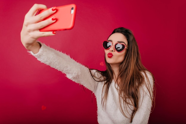 Chica guapa morena con suéter esponjoso y gafas de sol tomando una foto de sí misma. Maravilloso modelo femenino de pelo largo con corazón en la mejilla haciendo selfie con teléfono.