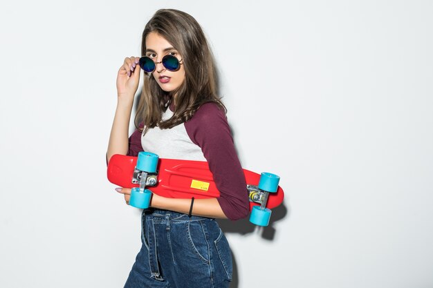 Chica guapa patinadora en ropa casual y gafas de sol negras con patineta roja aislada en la pared blanca