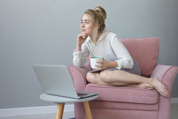 Chica guapa con peinado desordenado con expresión facial soñadora, disfrutando de un café, sentada en un cómodo sillón frente a una computadora portátil abierta, pensando en planes futuros y vacaciones de verano