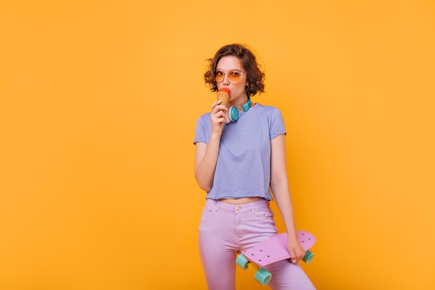 Chica guapa con pelo corto y oscuro comiendo helado. Foto interior de mujer espectacular complacida con patineta.