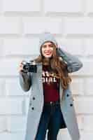 Foto gratuita chica guapa con pelo largo en abrigo gris en la pared al aire libre. ella sostiene la cámara, toca el gorro de punto y sonríe.