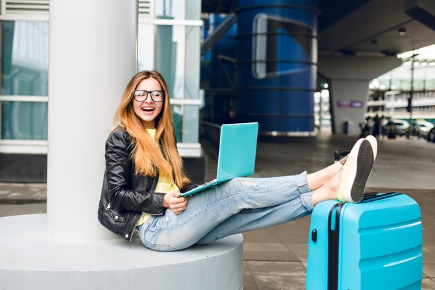 Chica guapa con pelo largo con gafas está sentada afuera en el aeropuerto. Viste jeans, chaqueta negra, zapatos amarillos y tiene laptop. Ella puso sus piernas en la maleta y sonrió a la cámara.