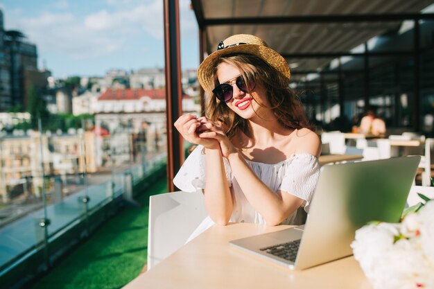 Chica guapa con pelo largo con gafas de sol se sienta a la mesa en la terraza de la cafetería. Lleva un vestido blanco con hombros descubiertos, lápiz labial rojo, sombrero. Ella está sonriendo a un lado.