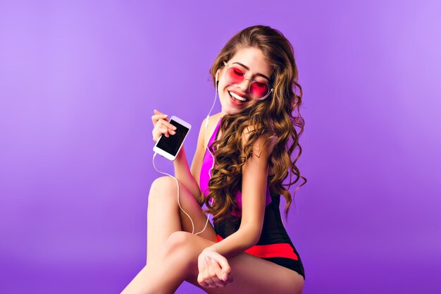 Chica guapa con pelo largo y rizado en gafas de sol rosas sobre fondo morado en estudio. Viste traje de baño y escucha música con auriculares.