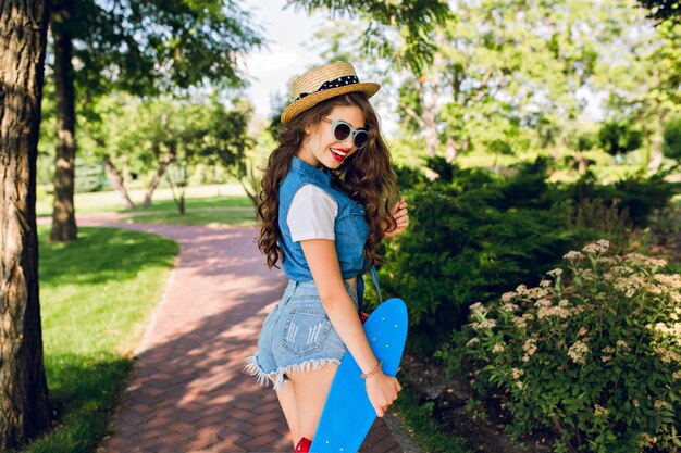 Chica guapa con pelo largo y rizado con sombrero y gafas de sol está caminando con patineta en el parque de verano. Lleva pantalones vaqueros, pantalones cortos. Ella está sonriendo con labios rojos.