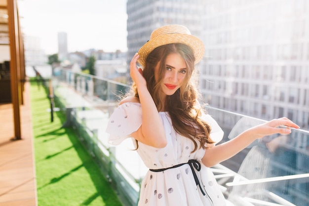 Chica guapa con pelo largo con sombrero está disfrutando en la terraza del balcón. Lleva un vestido blanco con hombros descubiertos y lápiz labial rojo. Ella está mirando a la cámara.