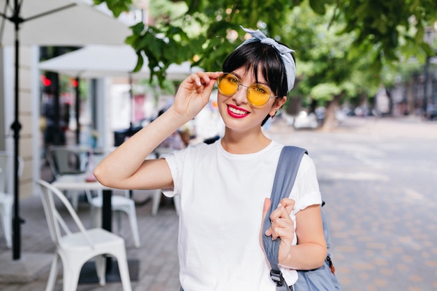 Chica guapa con pelo negro corto sonriendo y sosteniendo la mochila azul en un hombro mientras posa en la calle por la mañana