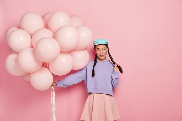 Chica guapa pensativa con mejillas rojas, vestida con ropa de moda, sostiene un montón de globos, viene de fiesta