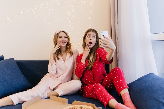 Chica guapa en pijama rojo y calcetines haciendo selfie con hermana y expresando asombro. Amigas positivas divirtiéndose mientras comen pizza.