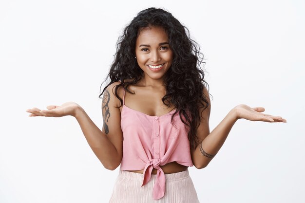 Chica guapa relajada con tatuajes, camiseta recortada, encogiéndose de hombros y manos extendidas hacia los lados despreocupada, de pie despistada, no se preocupe, exprese sentimientos despreocupados, pared blanca