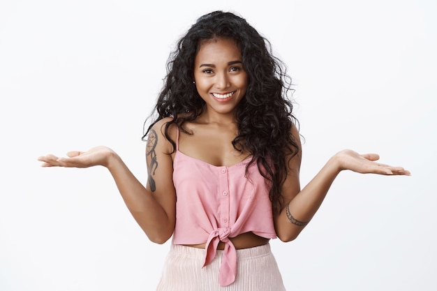 Chica guapa relajada con tatuajes, camiseta recortada, encogiéndose de hombros y manos extendidas hacia los lados despreocupada, de pie despistada, no se preocupe, exprese sentimientos despreocupados, pared blanca