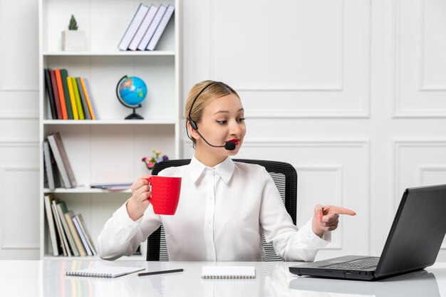Chica guapa rubia de servicio al cliente en camisa blanca con computadora portátil y auriculares apuntando en la pantalla
