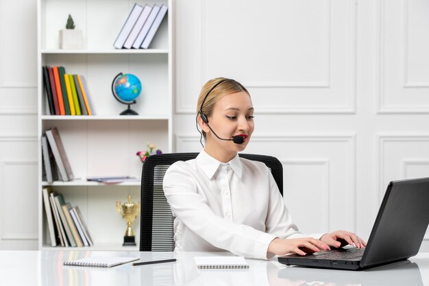 Chica guapa rubia de servicio al cliente en camisa blanca con computadora portátil y auriculares escribiendo en el teclado