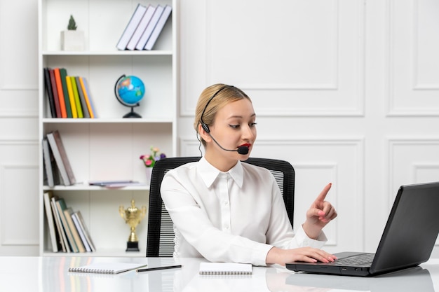 Chica guapa rubia de servicio al cliente con camisa blanca con computadora portátil y auriculares que muestran un minuto