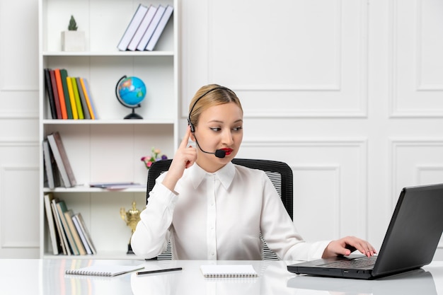 Chica guapa rubia de servicio al cliente con camisa blanca con laptop y auriculares hablando