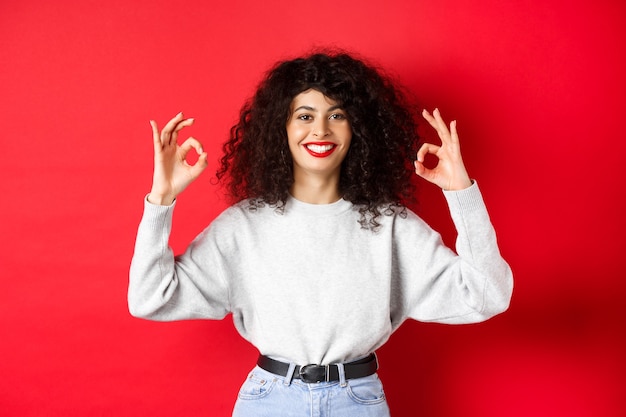 Chica guapa segura con peinado rizado, mostrando buenos gestos y sonriendo, aprueba y está de acuerdo con usted, alabando la excelente elección, de pie satisfecha sobre fondo rojo