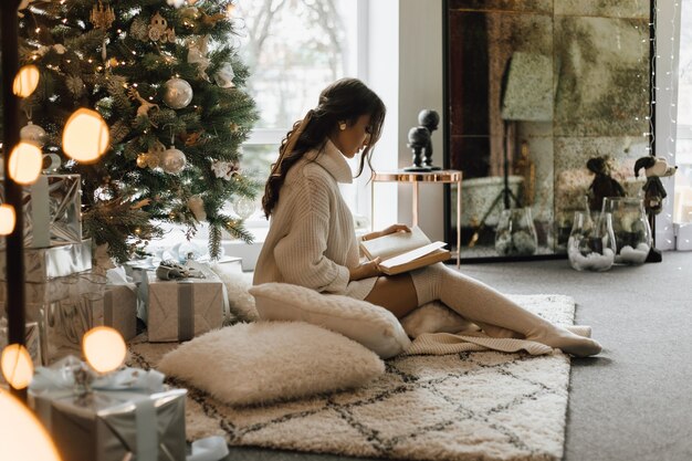 Chica guapa se sienta con almohadas y cuadros cerca de un árbol de Navidad y lee un libro