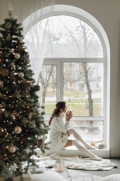 Chica guapa se sienta con una taza de té y cuadros cerca de un árbol de Navidad